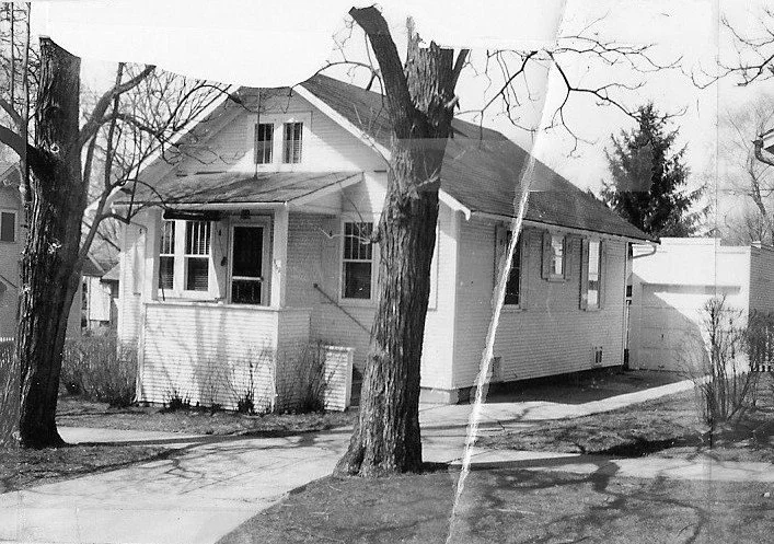 Black-and-white photo of a single-story bungalow with a covered front porch, two large trees in the yard, and a driveway leading to a detached garage.