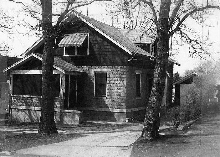 Black-and-white photo of a two-story house with a covered front porch, a striped window awning, two large leafless trees in front, and a driveway at the right.