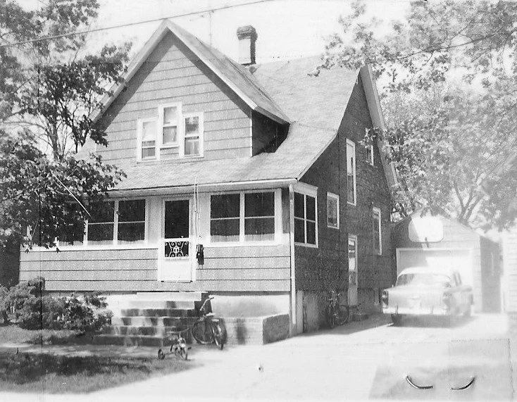 Black-and-white photo of a two-story house with an enclosed front porch, steps with a bicycle nearby, and a car parked in the driveway beside a garage.