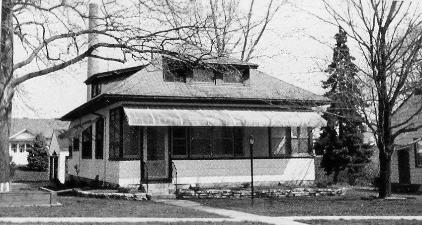 Black-and-white photo of a single-story bungalow with an enclosed front porch and awning, leafless trees and a small lamp post in the yard.