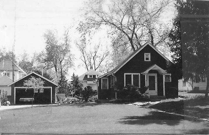 Black-and-white photo of a small bungalow-style house with a detached garage, driveway, and tall trees in the background.