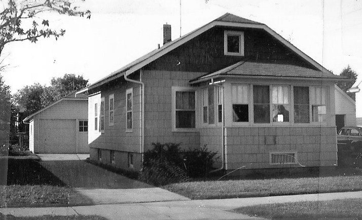 Black-and-white photo of a single-story bungalow with an enclosed front porch, driveway leading to a detached garage, front lawn and sidewalk.