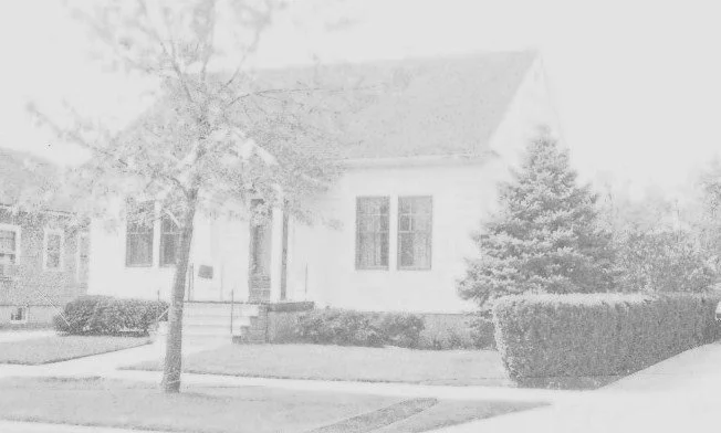 Single-story white bungalow with a front porch, a tree in the yard, and trimmed bushes along the sidewalk.