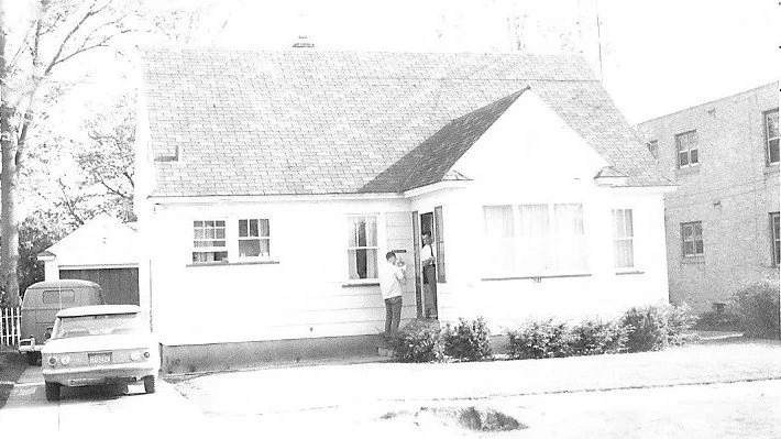 White single-story house with pitched roof; a person at the open front door, a car in the driveway, detached garage at left and an apartment building to the right.