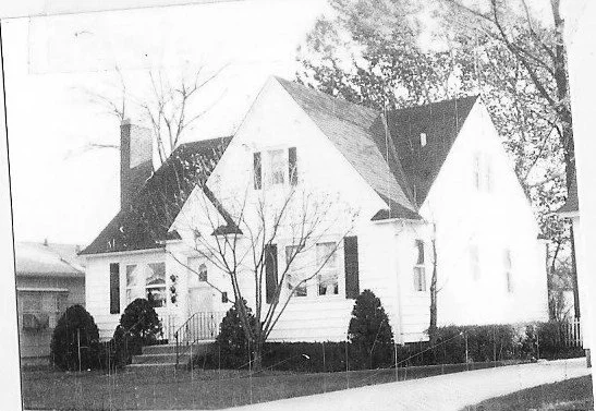 Black-and-white photo of a two-story gable-roof house with front steps, shrubs, a bare tree, and a sidewalk in front.
