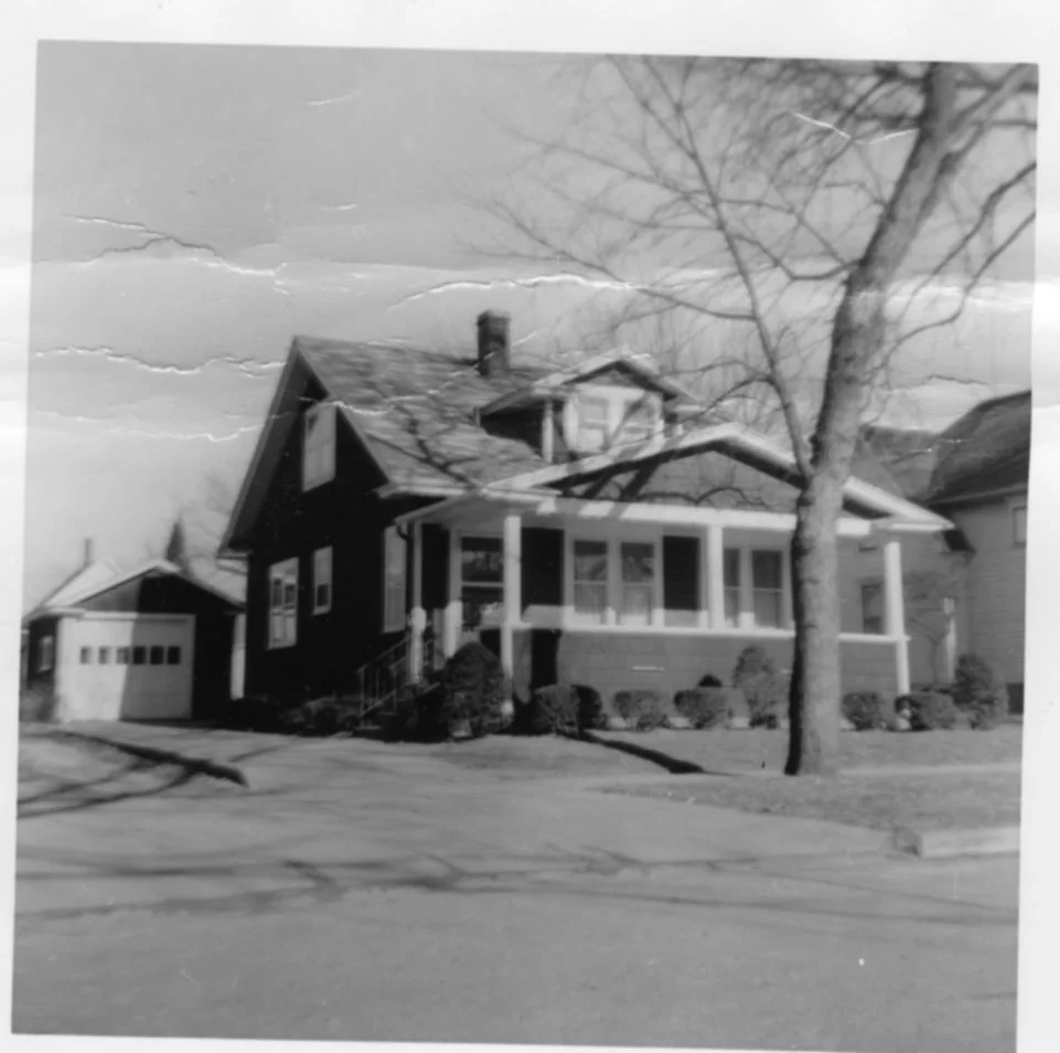 Black-and-white vintage photo of a two-story bungalow with a covered front porch, detached garage to the left, a leafless tree in the front yard, and visible creases on the print.
