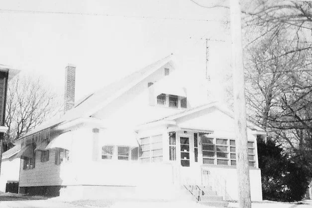 Front view of a white bungalow with an enclosed glass front porch, steps up to the door, a chimney, bare trees and a utility pole.