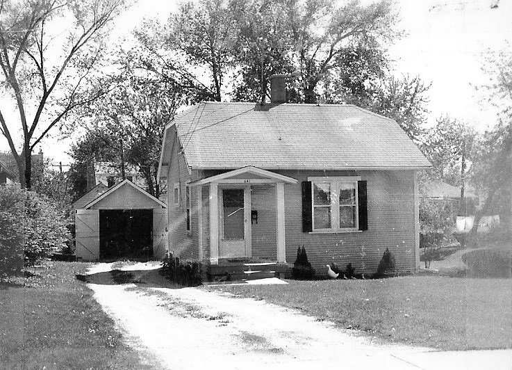 Black-and-white photo of a small one-story house with a front porch, detached garage, driveway, and large trees in the yard.