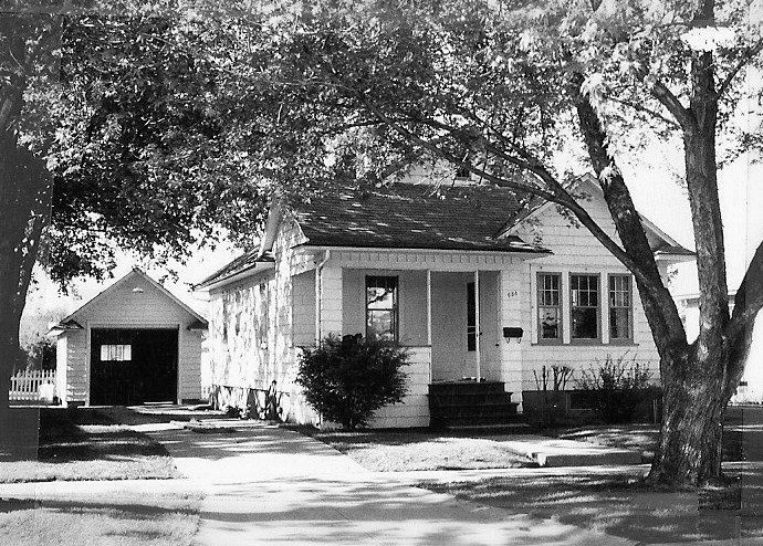 Black-and-white photo of a small single-story white house with a front porch and detached one-car garage, shaded by large trees.