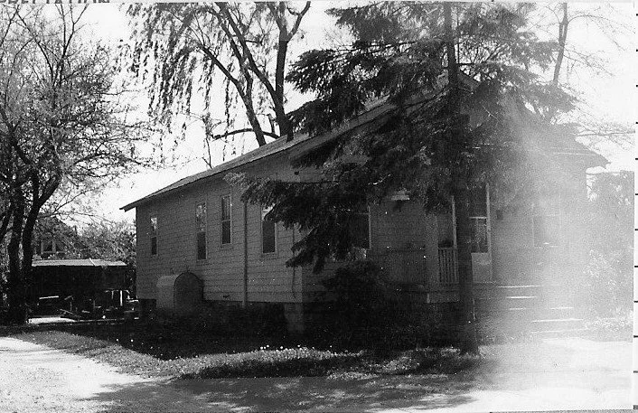 Black-and-white photo of a single-story wood-sided house partly shaded by large trees, with a small front porch and a driveway to the left.