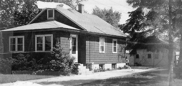 Black-and-white photo of a single-story wood-sided bungalow with an enclosed front porch, driveway to a detached garage, and trees in the yard.