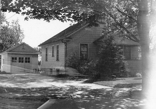 Black-and-white photo of a small single-story house with a detached garage and large trees casting shadows across the yard and driveway.