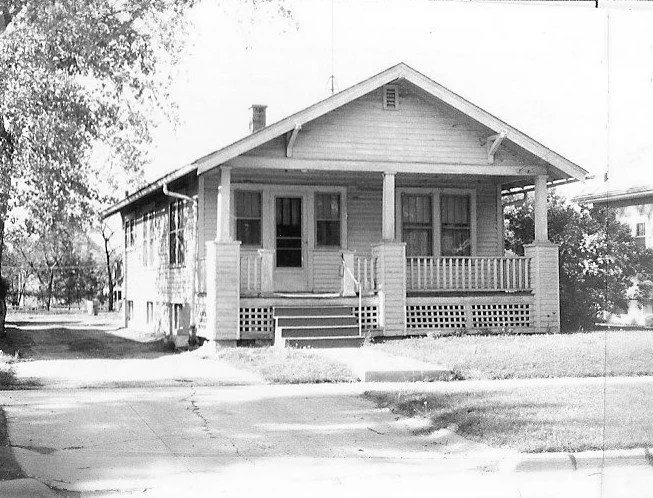 Black-and-white photo of a single-story bungalow with a covered front porch, square columns, steps, and a small yard facing the sidewalk.