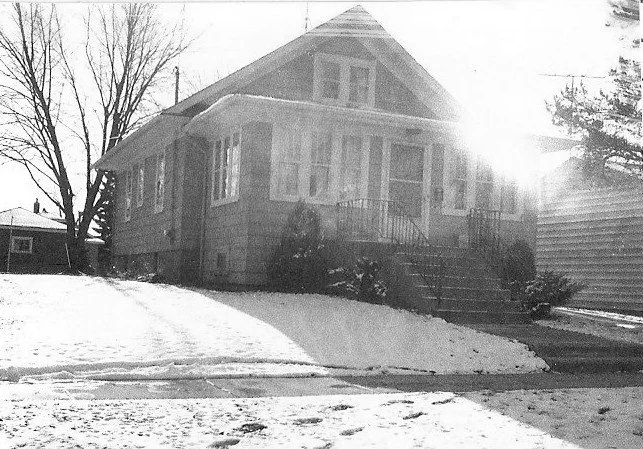 Black-and-white photo of a single-story bungalow with a snowy front yard, front steps and railing, and bright sunlight glare.