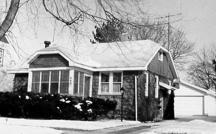 Black-and-white photo of a small bungalow with snow on the ground, enclosed front porch (house number 680), bare trees, and a detached garage.