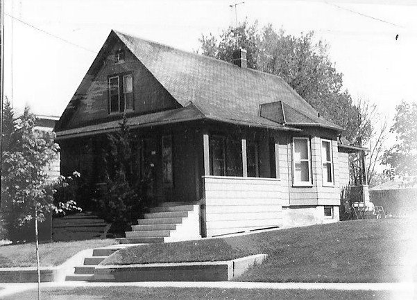 Black-and-white photo of a single-story gable-roof bungalow with front steps, enclosed porch, lawn, and trees.