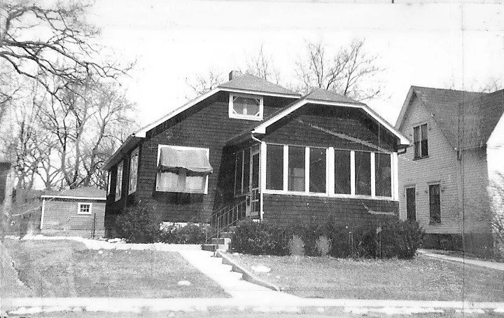 Black-and-white photo of a single-story bungalow with an enclosed front porch, concrete walkway, shrubs, and a neighboring house.