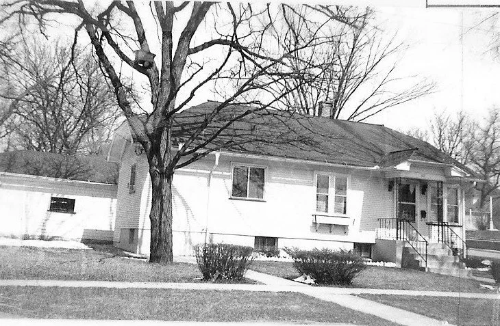 Black-and-white photo of a single-story brick house with a small front porch and steps, a large leafless tree in the yard, low shrubs, and a detached garage.