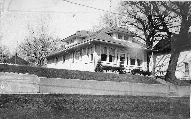Black-and-white photo of a single-story bungalow atop a raised lawn with a concrete retaining wall and leafless trees.
