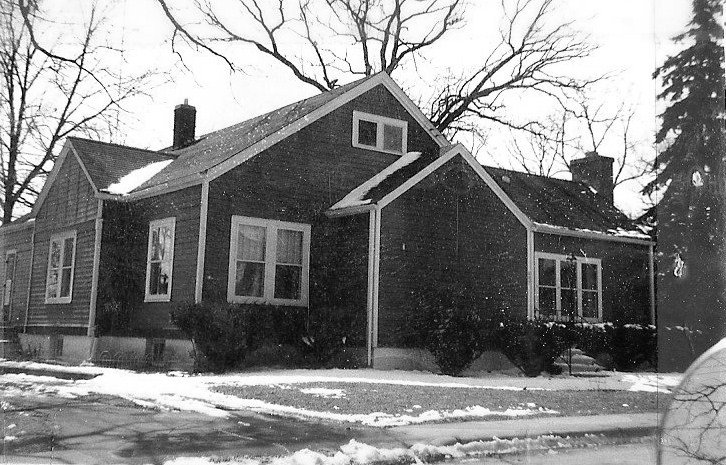 Black-and-white photo of a single-story gabled house (bungalow) with snow on the ground and bare trees in the background.