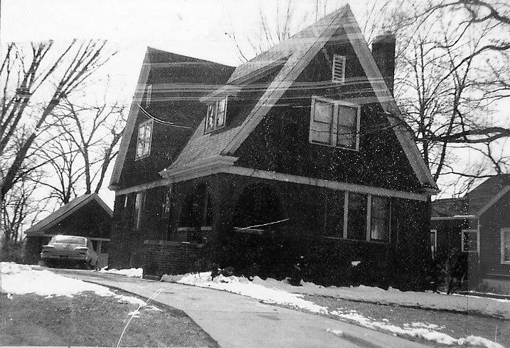 Black-and-white photo of a two-story, steep‑roof house with a front porch, snow along the driveway and a car parked by a detached garage.