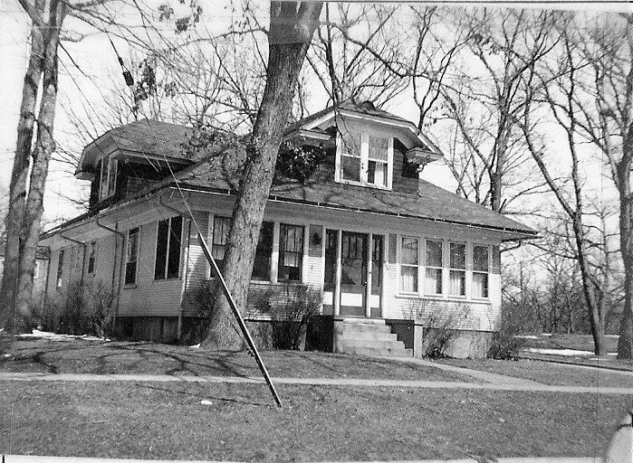 Black-and-white photo of a two-story Craftsman-style house with an enclosed front porch, leafless trees surrounding it and a slightly leaning utility pole in the front yard.