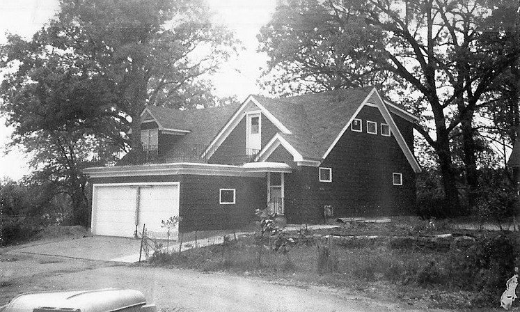 Black-and-white photo of a two-story gabled house with an attached two-car garage, large trees around it, an overgrown front yard, and part of a vintage car visible in the foreground.