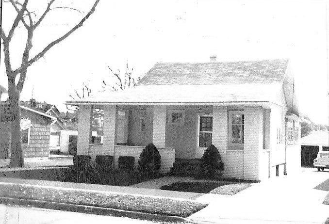 Black-and-white photo of a small single-story bungalow with a covered front porch, shrubs, a bare tree at left, and a car in the driveway.