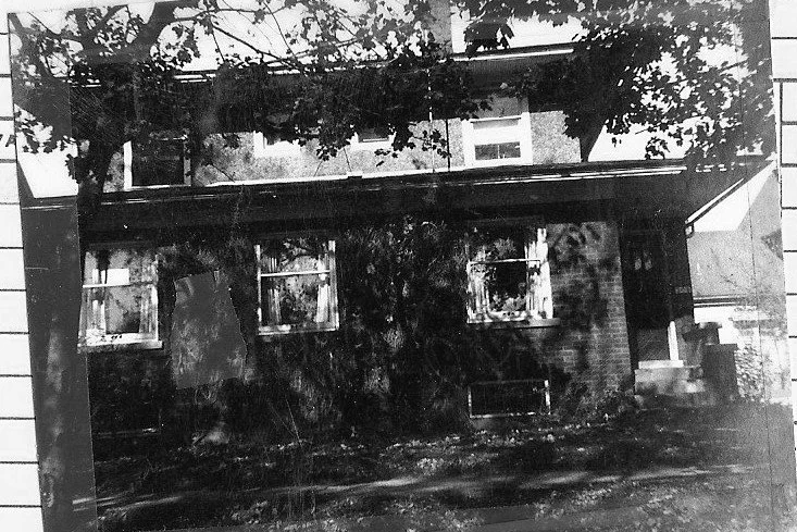 Black-and-white photo of a two-story brick house with three front windows, a small porch and steps on the right, and tree branches casting shadows across the facade.