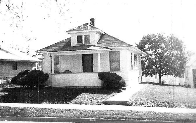 Black-and-white photo of a small one-story bungalow with a covered front porch, two large shrubs, a sidewalk leading to the entrance, and a tree on the right.