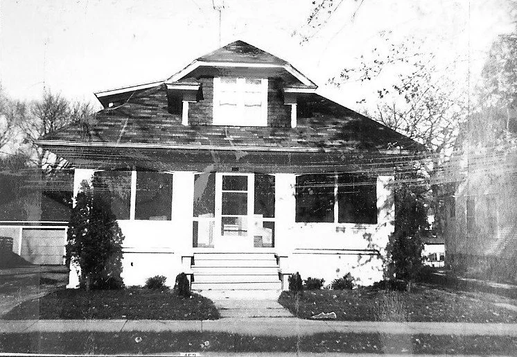 Black-and-white photo of a single-story bungalow with a covered front porch, central steps, dormer window, and shrubs on the lawn.
