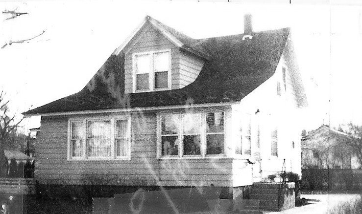 Black-and-white photo of an older two-story wood-frame house with an enclosed front porch, dormer windows, and a pitched roof.