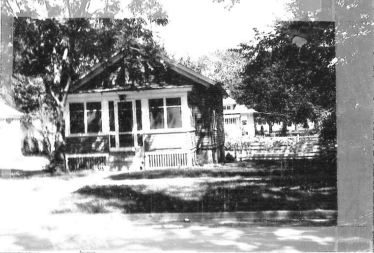 Black-and-white vintage photo of a small single-story bungalow with a screened front porch, large shade trees, and a grassy yard.