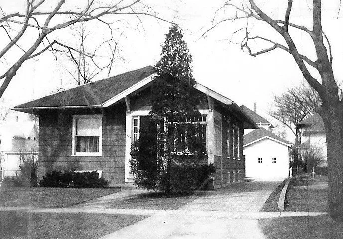Black-and-white photo of a single-story bungalow with an enclosed front porch, driveway leading to a detached garage, and bare trees.