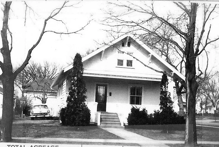 Black-and-white photo of a small front-porch bungalow surrounded by shrubs and leafless trees, with a vintage car parked to the left.