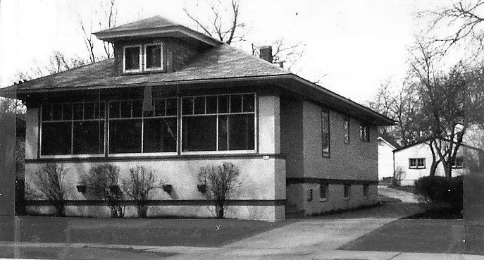 Black-and-white photo of a single-story bungalow with an enclosed front porch and dormer window, driveway to the right and leafless trees in the background.
