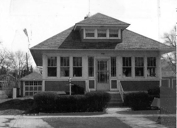 Black-and-white photo of a one-story bungalow with a central dormer, wide enclosed front porch, trimmed shrubs, and a detached garage.