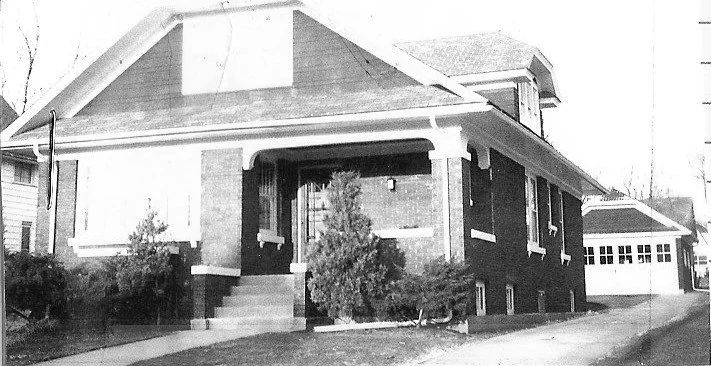 Black-and-white photo of a one-story brick bungalow with a covered front porch, small shrubs, and a detached garage at the side.