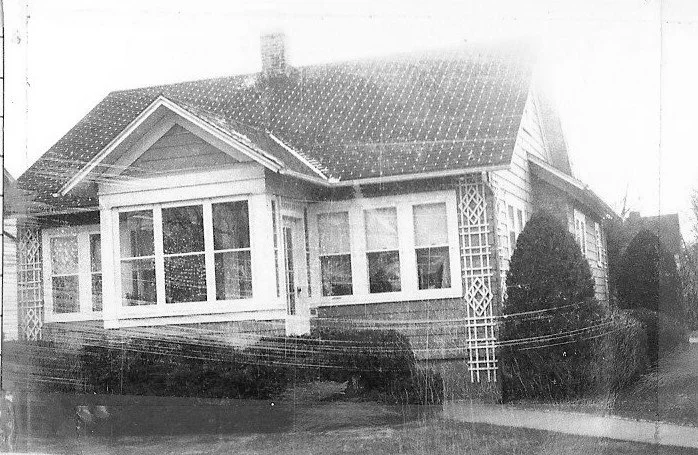 Black-and-white photo of a single-story bungalow with an enclosed front porch, lattice columns, shrubs along the foundation, and a sloped roof with a chimney.