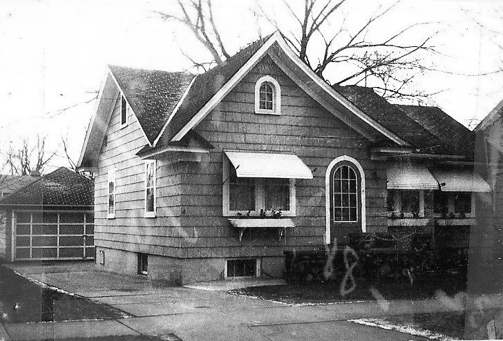 Black-and-white photo of a small gabled bungalow with awnings over the front windows, a curved-pane entry door, and a detached garage in the background.