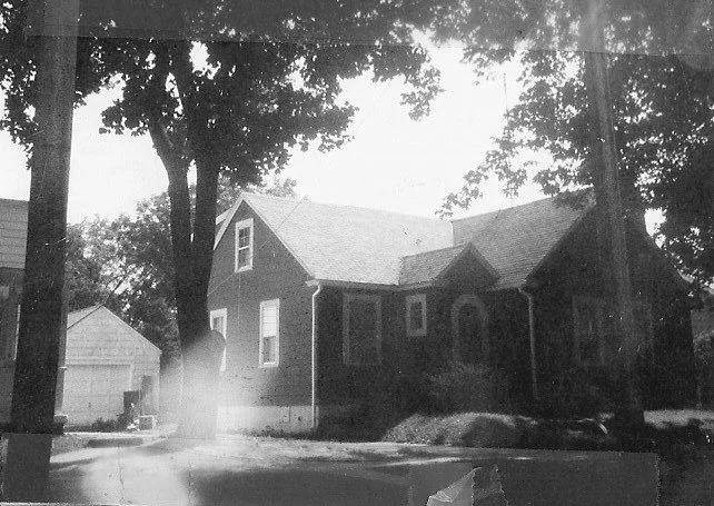 Black-and-white photo of a one-story brick suburban house with tall trees and a detached garage to the left.