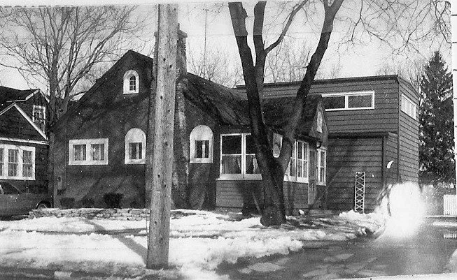 Black-and-white photo of a small house with snow-covered front yard, bare trees and a utility pole in the foreground.