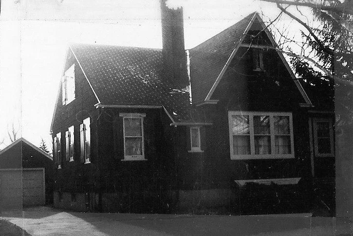 Black-and-white photo of a two-story gabled house with a tall chimney, front windows, driveway and detached garage.