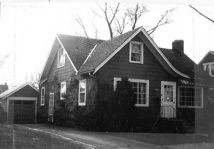 Black-and-white photo of a two-story bungalow-style house with a pitched roof, front porch, detached garage and a driveway, framed by leafless trees.