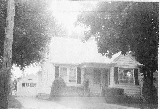 Black-and-white photo of a small single-story house with a front porch, shrubs, a driveway leading to a detached garage, and large trees framing the scene.