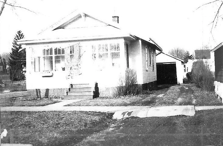 Black-and-white photo of a single-story bungalow with an enclosed front porch, concrete steps, detached garage to the right, and a lawn and sidewalk in the foreground.
