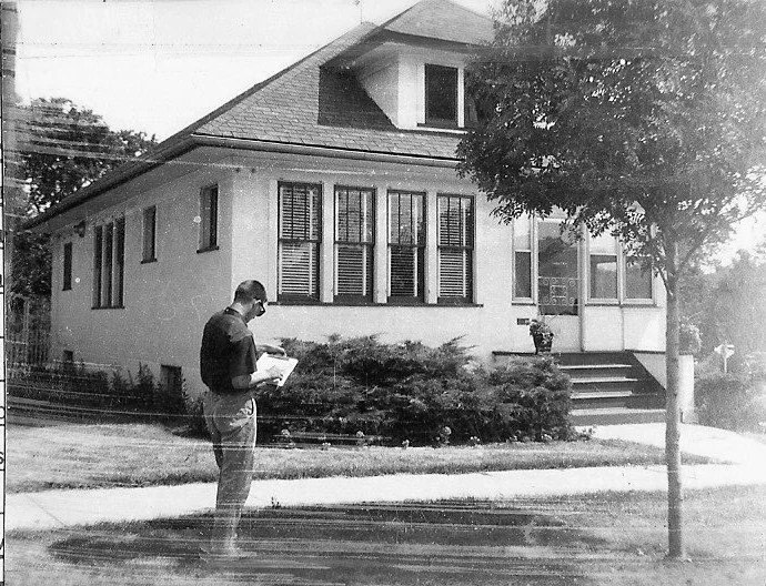 Black-and-white photo of a man standing on a sidewalk reading papers in front of a single-story house with shuttered windows, front steps, and a tree.