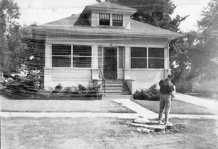Black-and-white photo of a single-story bungalow with an enclosed front porch and a man standing on the lawn near the sidewalk.