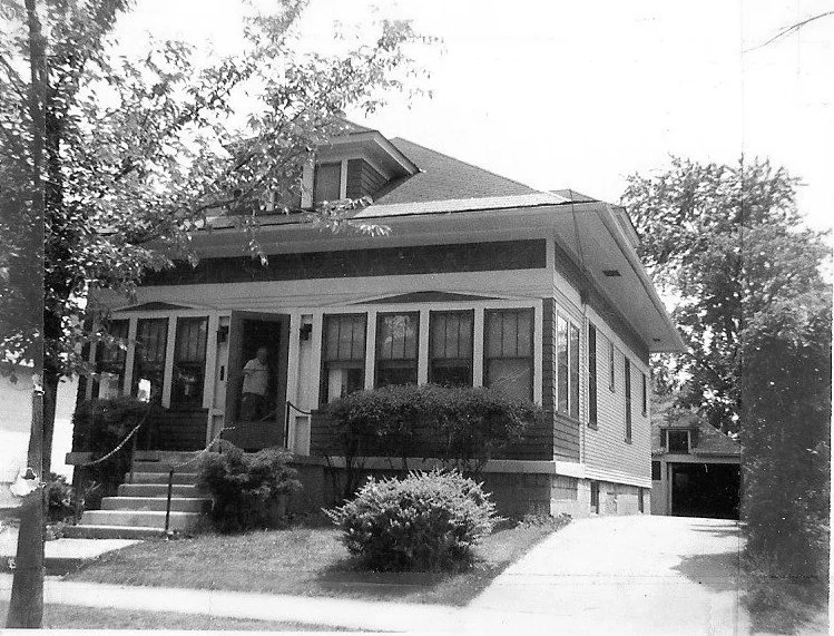 Black-and-white photo of a single-story bungalow with an enclosed front porch, steps and shrubs, a driveway to the right, and a person standing in the doorway.