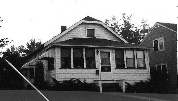 Black-and-white photo of a small bungalow with an enclosed front porch, siding, and a neighboring house to the right.
